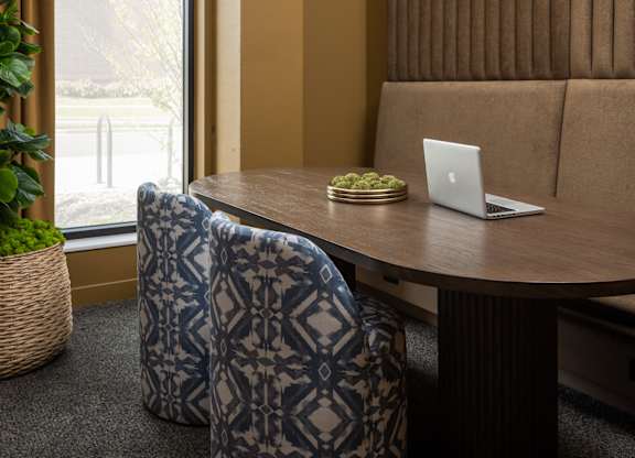 A room with a brown couch, a table with a laptop and a plant on it, and two patterned chairs.
