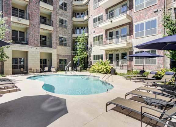 A swimming pool surrounded by lounge chairs and umbrellas in front of a brick building.