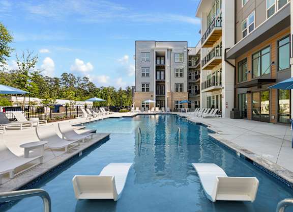 A swimming pool with white lounge chairs and apartment buildings in the background.