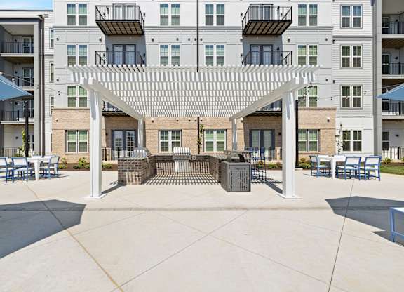 A patio with a white pergola and blue chairs is surrounded by apartment buildings.