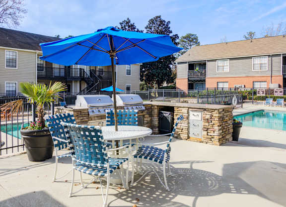 A blue umbrella shades a table with chairs and a potted plant.