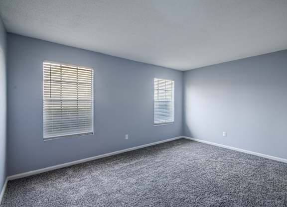 A room with grey carpet and two windows with blinds.