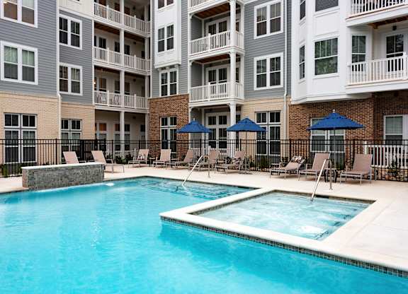 A swimming pool surrounded by chairs and umbrellas in front of apartment buildings.