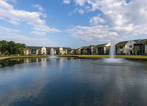 A fountain in the middle of a lake in front of apartment buildings.