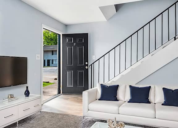A white couch with blue pillows sits in a living room with a television and a staircase.