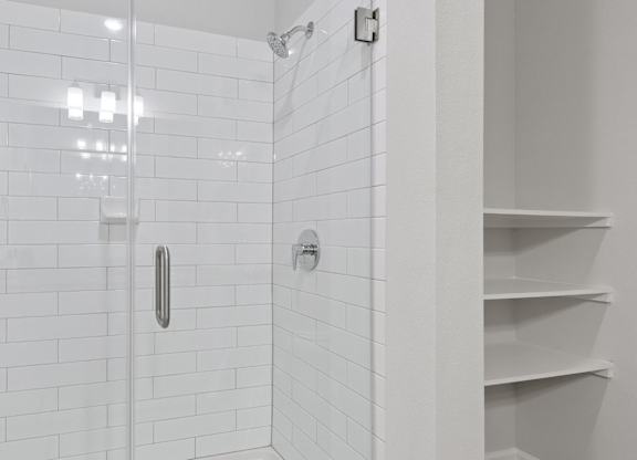 A white tiled shower with a glass door and a white shelf.
