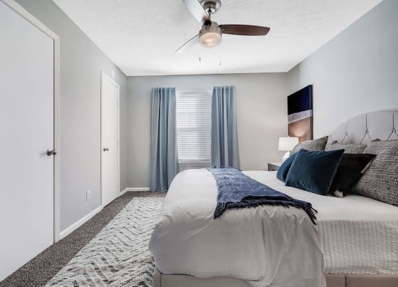 Apartments In Louisville, KY With In-Unit Washer And Dryer - Enclave At Breckenridge - Spacious Bedroom With Carpet Flooring, Large Bed, Nightstand with Lamp, Ceiling Fan, And Window.