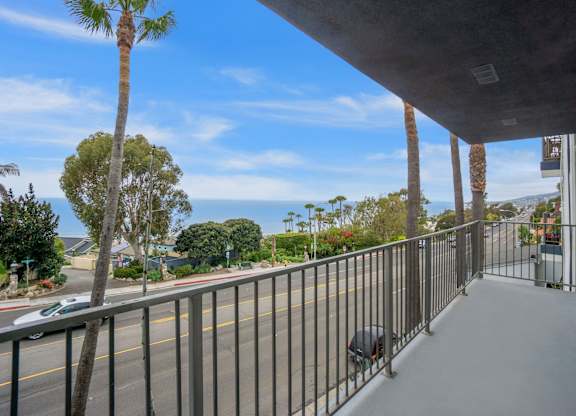 A balcony with a metal railing overlooks a street with cars and palm trees.