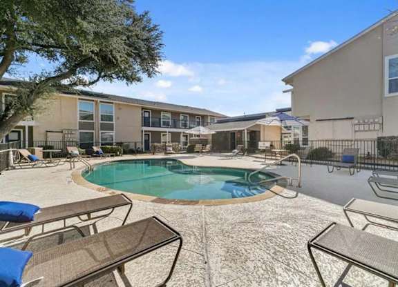 A pool surrounded by chairs and umbrellas in front of apartment buildings.