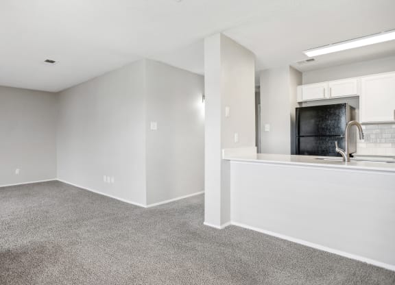 the living room and kitchen of an apartment with white walls and carpet