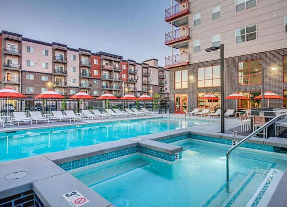 A swimming pool surrounded by lounge chairs and umbrellas in front of apartment buildings.