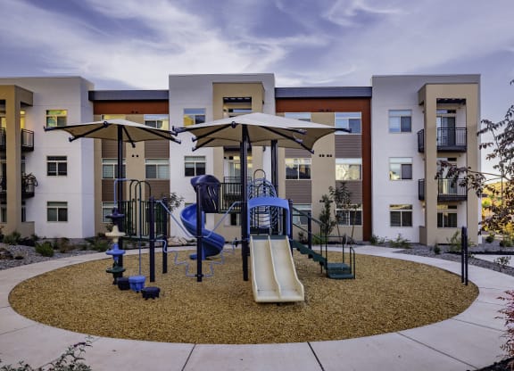 an outdoor playground with swings and umbrellas in front of apartments