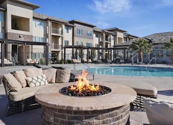 A fire pit sits in the middle of a patio with a pool and apartment buildings in the background.