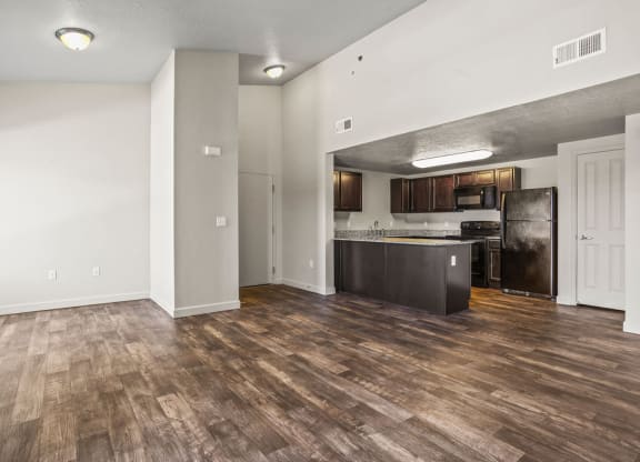 an empty living room with a kitchen with a refrigerator