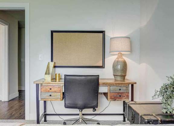 A black chair sits in front of a desk with a lamp and a picture frame on it.