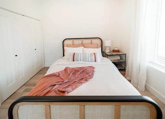 A bed with a white sheet and a pink blanket is in the foreground of a room with a white door and a nightstand with a lamp.