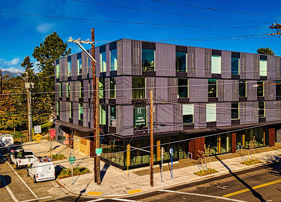 A modern building with a curved facade and large windows is surrounded by a parking lot and trees.