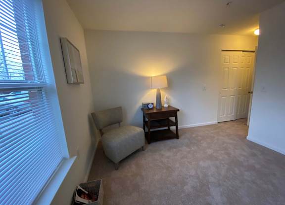 A living room with a chair, a table with a lamp and a clock on it, and a window with blinds.