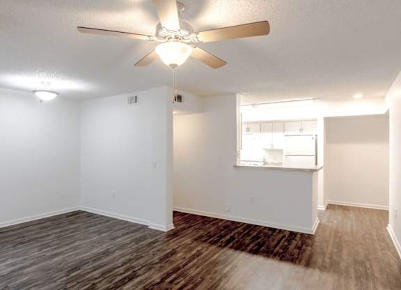 an empty living room with a ceiling fan and a kitchen in the background