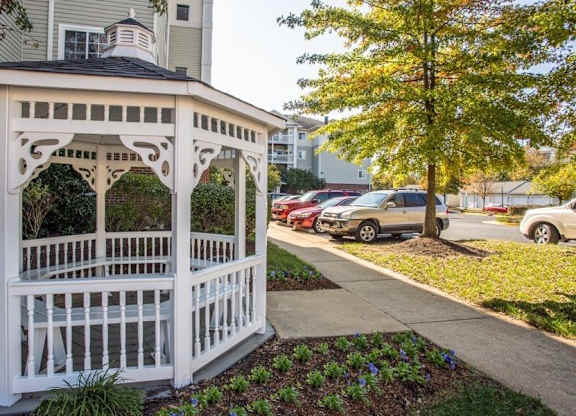 A white gazebo is situated in a landscaped area in front of a row of parked cars.