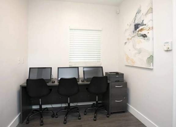 Three computers are on a desk with chairs.