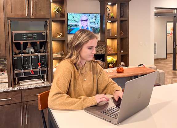 A woman in a yellow sweater is working on a laptop in a modern kitchen.