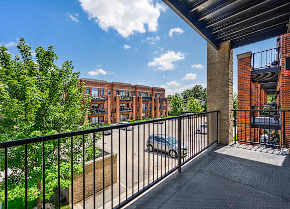 A balcony with a black railing overlooks a parking lot and a building.