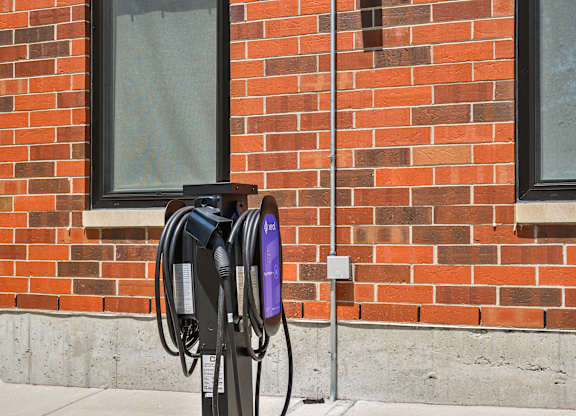 A black electric vehicle charging station is installed on a sidewalk in front of a red brick building.