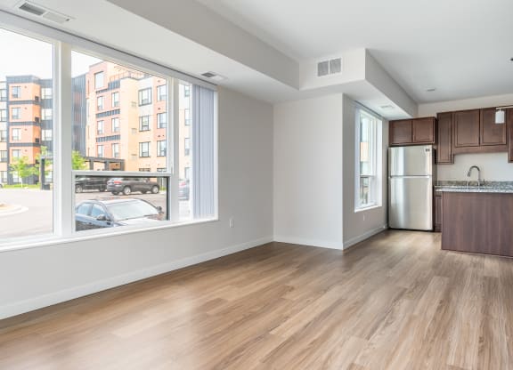 an empty living room with a large window and a kitchen in the background