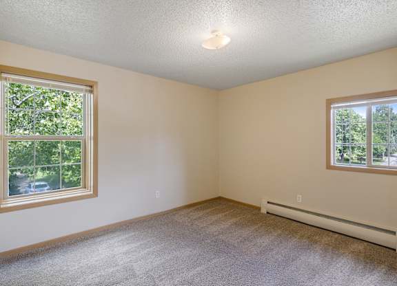 A room with a carpeted floor, a window with a view of trees, and a wall-mounted air conditioner.