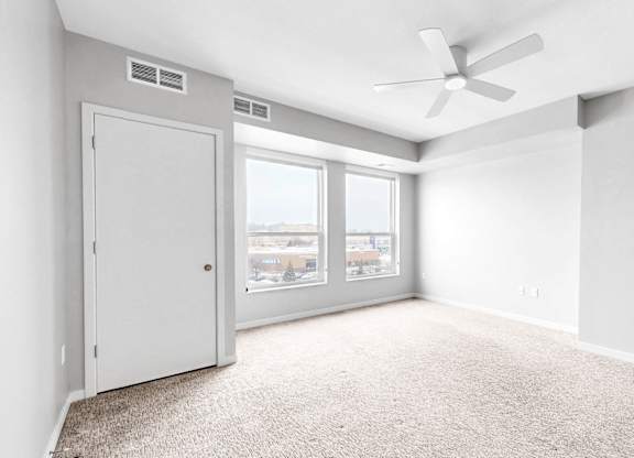 A room with a carpet floor, a ceiling fan, and a window with a view of buildings outside.