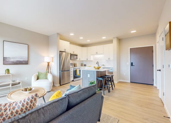 a living room with a couch a table and a chair and a kitchen with a refrigerator and