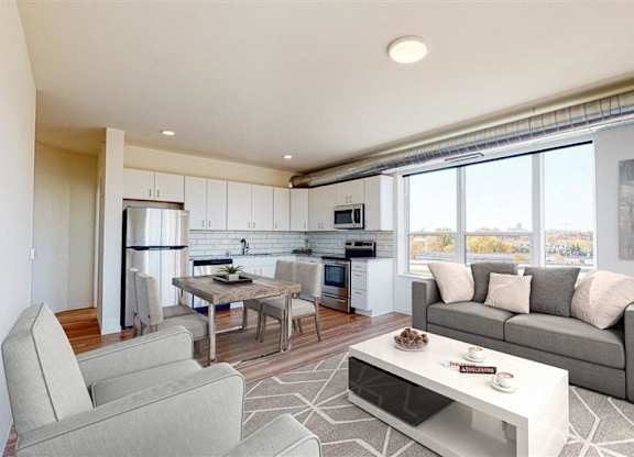 A modern living room with a grey couch, a white coffee table, and a kitchen area in the background at The Bessemer at Seward Commons Apartments, Minneapolis, MN,  55404