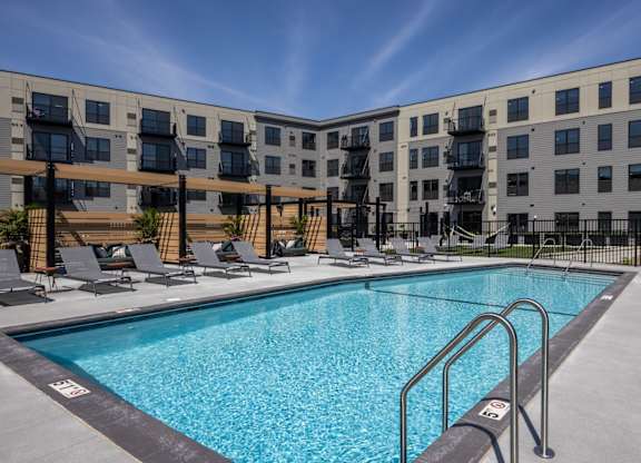 A swimming pool in front of a building with lounge chairs around it.