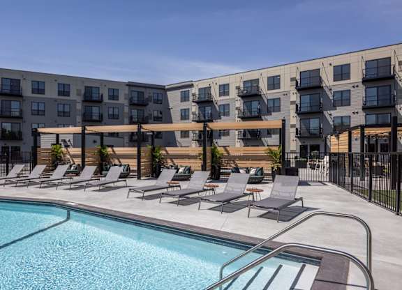 A swimming pool is surrounded by lounge chairs in front of apartment buildings.