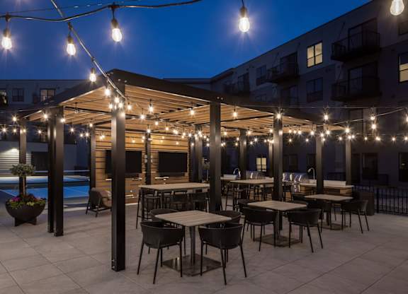A patio with tables and chairs is illuminated by string lights.