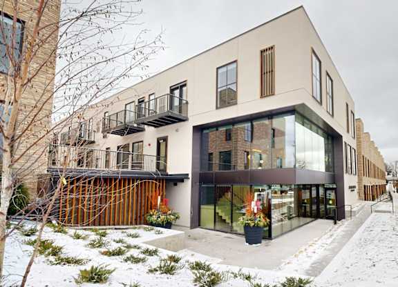 A modern building with a mix of beige and black exterior walls, large windows, and a balcony with a railing.