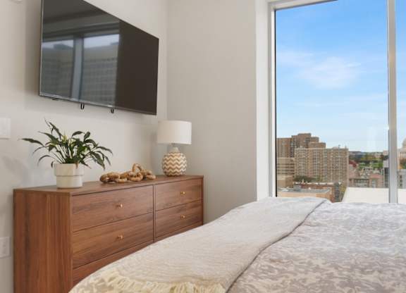 A bedroom with a large bed, a wooden dresser, and a view of the city through a large window.