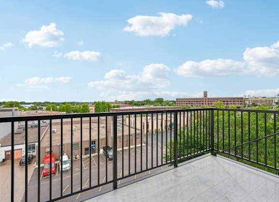 A balcony with a black railing overlooks a parking lot and buildings.