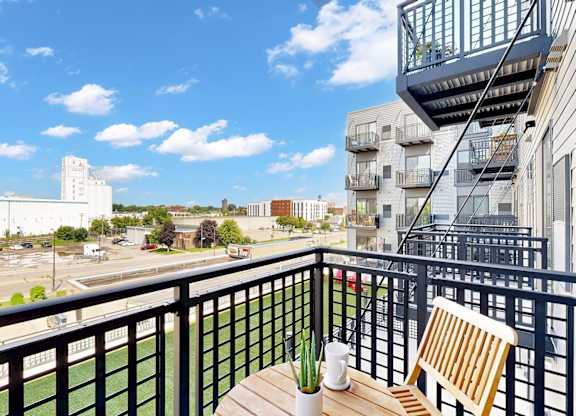 A balcony with a chair and table overlooking a parking lot and buildings.