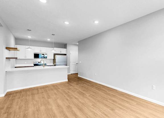 A large empty room with wooden flooring and a kitchen area in the background.