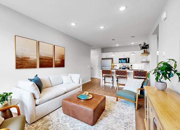 A living room with a white couch, a brown ottoman, and a TV in the background.