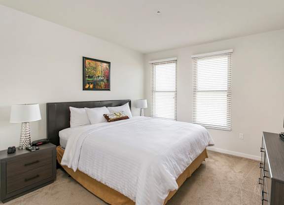 A bedroom with a large bed, a nightstand with a lamp, and a painting on the wall. at Cirrus Redwood City Apartments, Redwood City, California