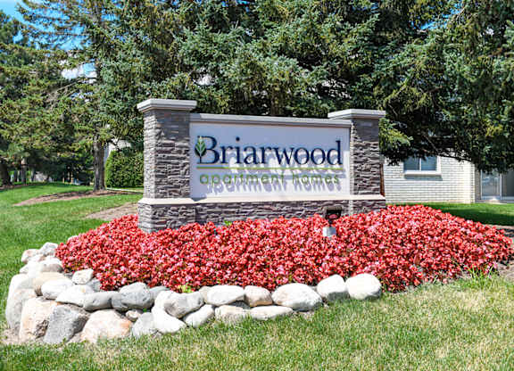 A sign for Briarwood Apartment Homes is surrounded by red flowers and rocks.