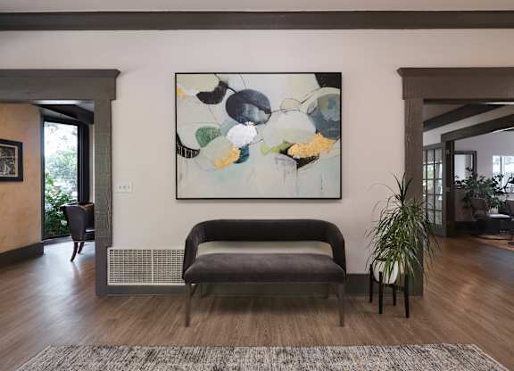 A large abstract painting hangs on a wall above a grey bench in a spacious room.