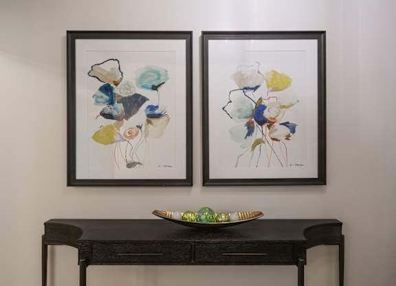 Two framed artworks hang on a wall above a dark desk with a green bowl on it.