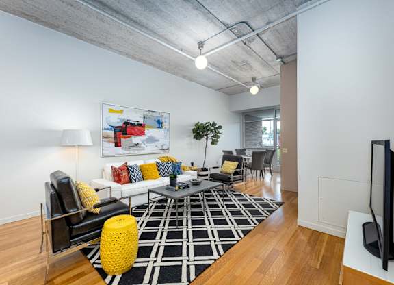 A living room with a black and white rug, a yellow pouffe, a black chair, a white couch with colorful pillows, a small tree, and a painting on the wall.