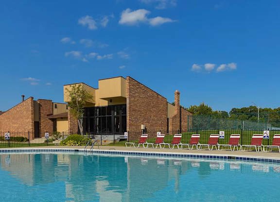 A swimming pool in front of a building with red chairs around it.
