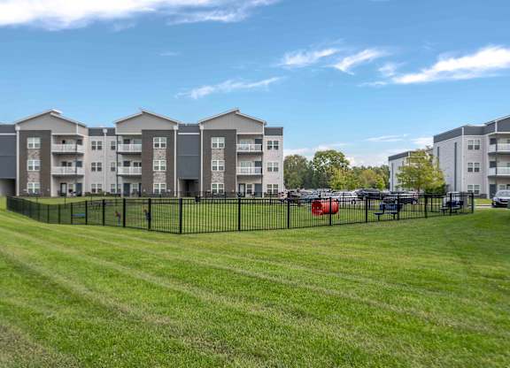 A row of apartment buildings with a grassy field in front and cars parked in the background.