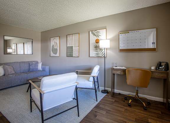 A living room with a grey couch, a white chair, a desk with a chair, a lamp, a calendar, and a mirror.